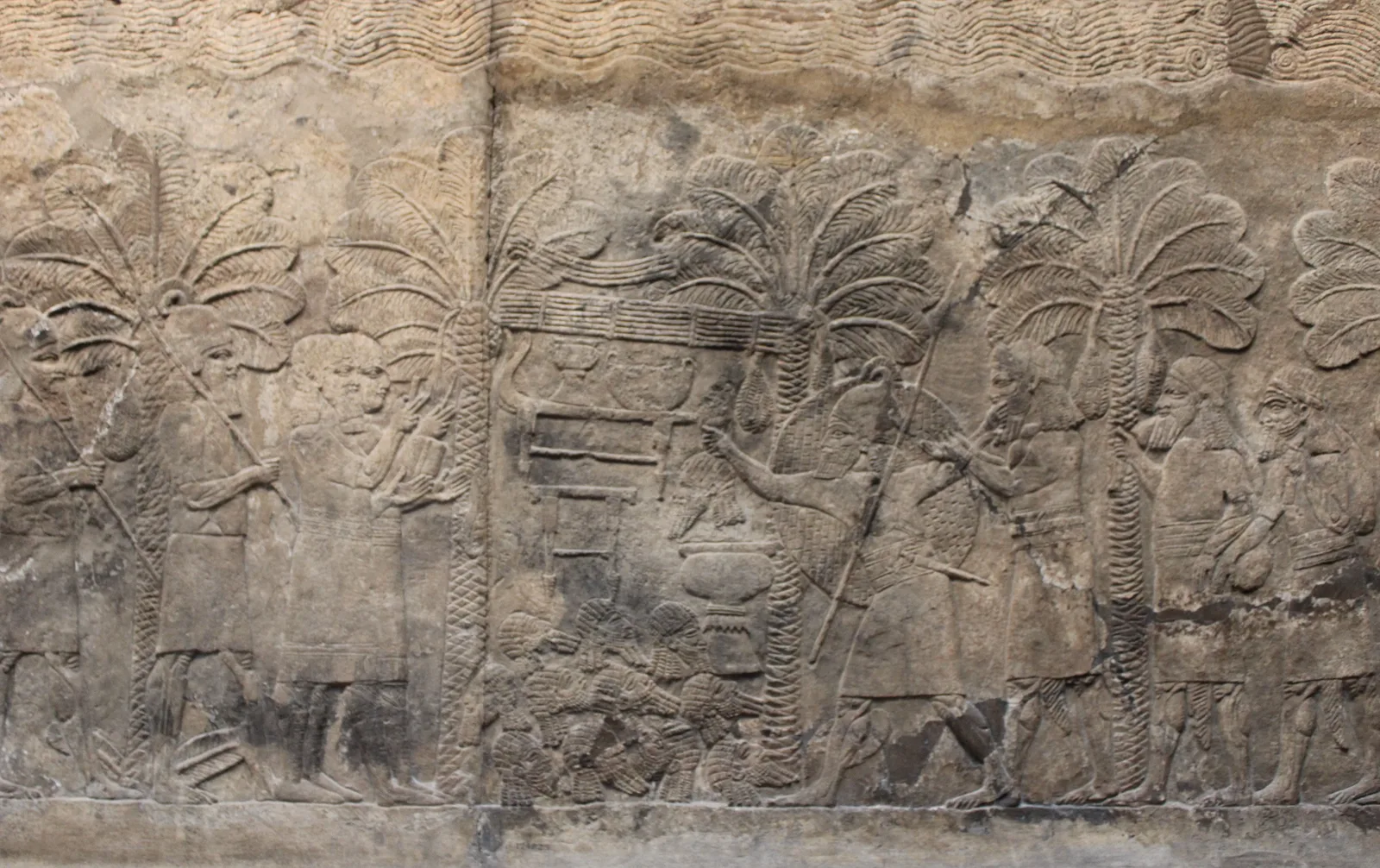 Relief from Nineveh: two Assyrian scribes, one holding a writing board, record booty beside soldiers under palm trees